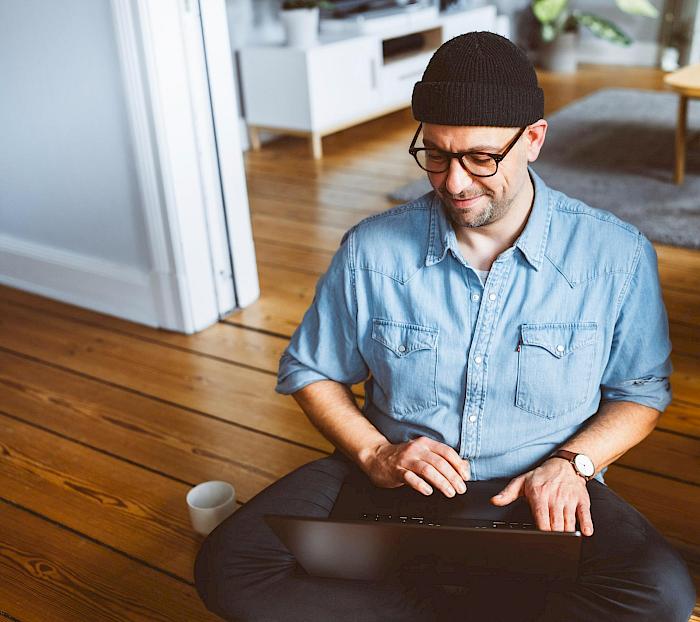 Ein mittelalter Mann sitzt im Schneidersitz auf einem Dielenboden. Auf dem Schoß liegt ein Laptop. Er blickt lächelnd auf den Bildschirm und tippt auf der Tastatur.